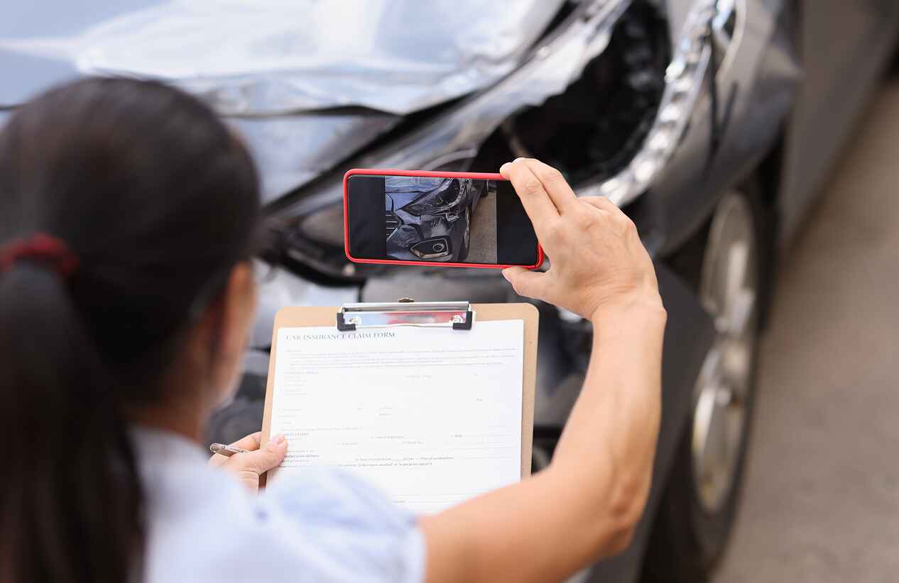 person performing inspection on wrecked car and documenting with photos while holding a clipboard