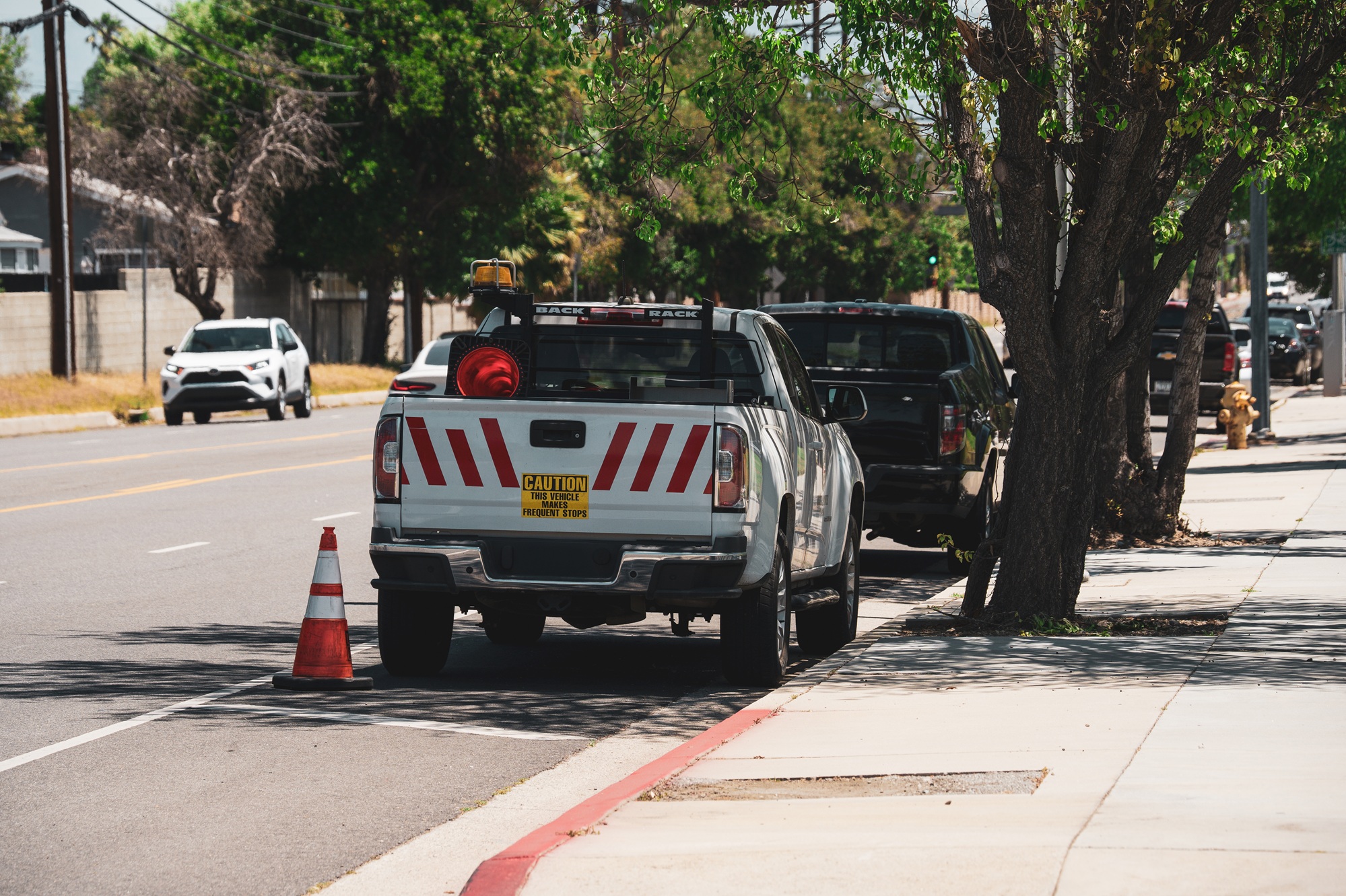 A white pickup truck with reflective safety stripes and caution decals is parked on the side of a road next to a sidewalk and trees
